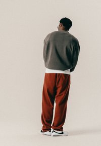 Άνδρας με γκρι φούτερ, λευκό εσώρουχο, κόκκινες παντελόνες και λευκά αθλητικά παπούτσια στέκεται με την πλάτη στραμμένη προς το κοινό σε έναν απλό ανοιχτό φόντο.