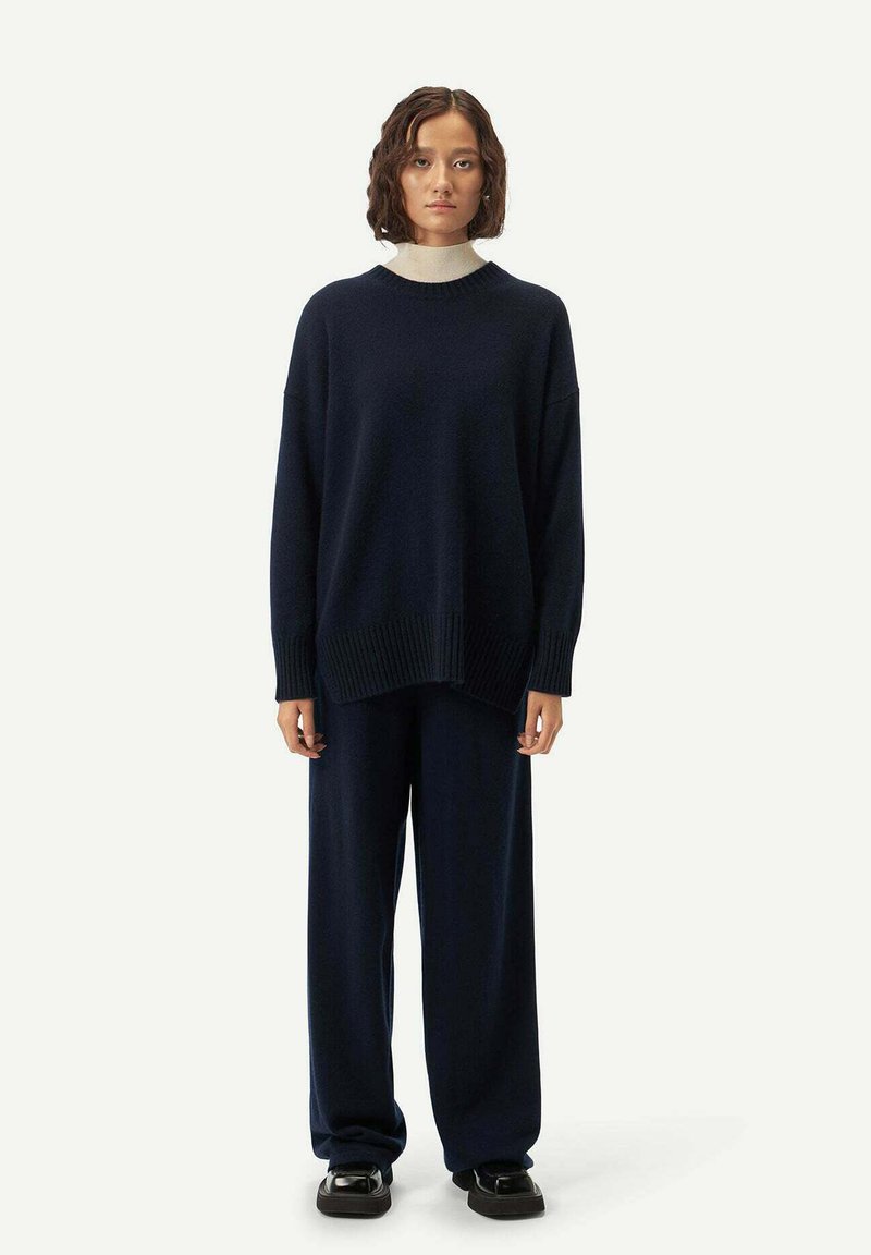 Marinblå oversized stickad tröja med ribbad nederkant, matchad med vida byxor och svarta skor, mot en neutral bakgrund.
