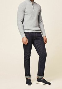 Uomo che indossa un maglione zip grigio chiaro, jeans scuri arrotolati e scarpe nere con suole bianche, in piedi con uno sfondo neutro.