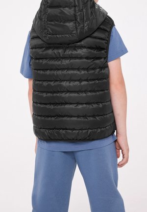 Kind trägt eine schwarze Kapuzen-Steppweste über einem blauen kurzärmeligen Hemd und blauen Hosen, steht mit dem Rücken zur Kamera vor einem weißen Hintergrund.