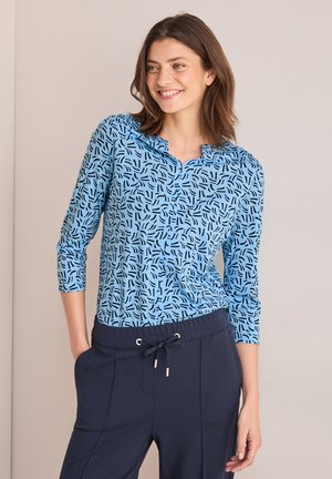 Blauw top met een onderscheidend zwart abstract patroon, driekwart mouwen en een gegolfde halslijn, gecombineerd met donkere marineblauwe broek.