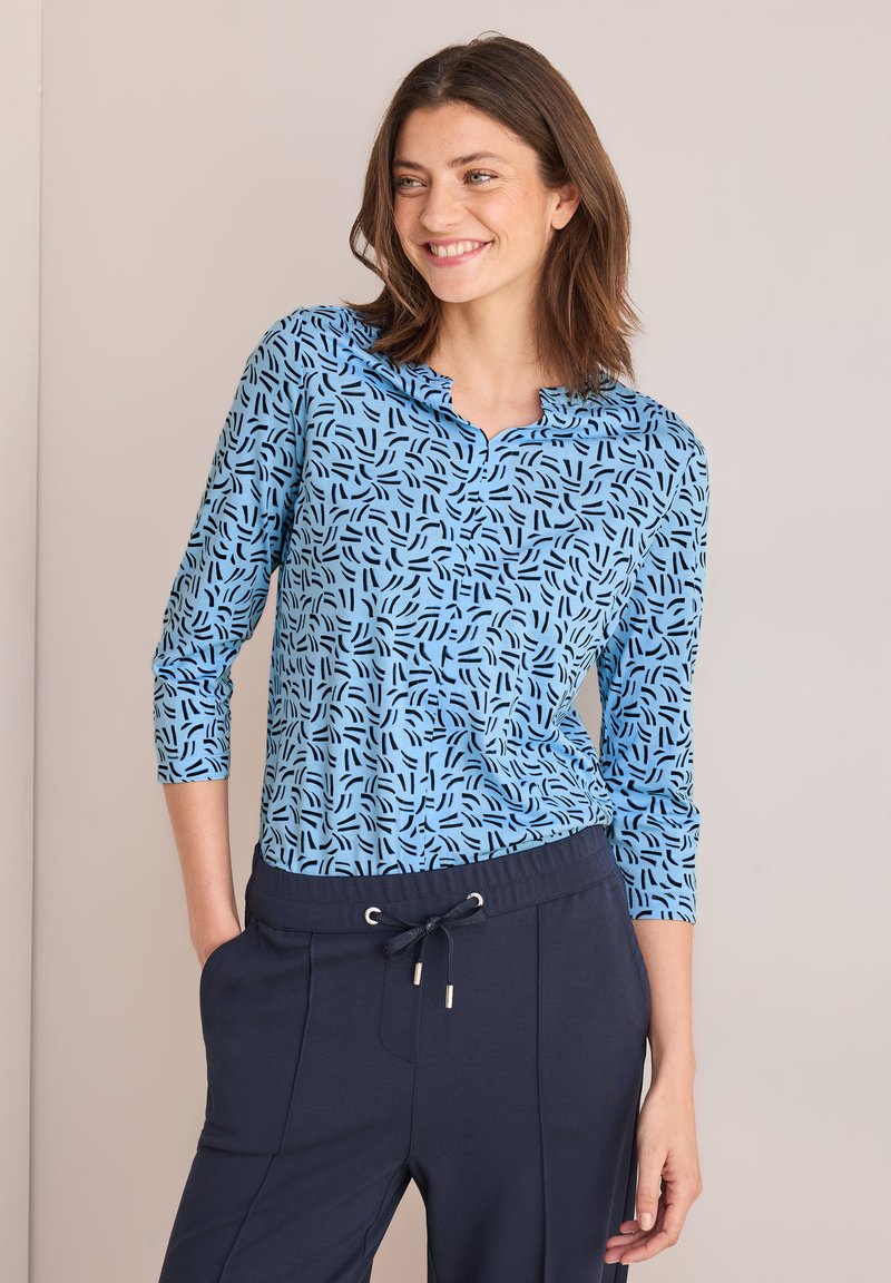 Blaue Bluse mit einem markanten schwarzen abstrakten Muster, dreiviertellangen Ärmeln und einem gewellten Ausschnitt, kombiniert mit dunkelblauen Hosen.