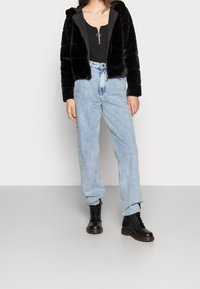 Chaqueta con capucha de piel sintética negra con superficie texturizada, usada sobre una camiseta negra. Vaqueros de tiro alto color azul claro y botas negras con cordones completan el conjunto.