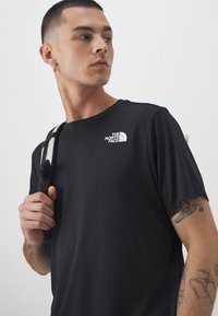 T-shirt preto de manga curta feito de um tecido macio, com um logo branco no lado esquerdo do peito. Uma alça de ombro é visível ao fundo.