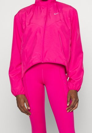 Tummaihoinen henkilö, jolla on kirkkaan pinkki Nike vetoketjutakki ja vastaavat pinkit legginssit yksiväristä vaaleaa taustaa vasten.