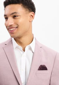 Lichtroze blazer met een subtiele textuur, scherpe revers en een borstzak met een donker bordeaux rode pochet over een wit shirt.