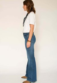 Femme debout de profil portant une chemise blanche à manches courtes, un jean bleu taille haute évasé, des bracelets bruns et des chaussures pointues couleur beige.