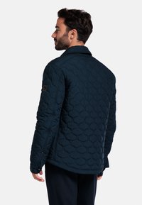 Marineblauwe gewatteerde jas met een gestructureerd patroon, kraag en zwarte hardwareaccenten. Heeft lange mouwen en een rechte zoom.