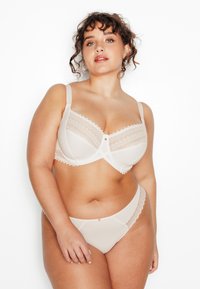 Ensemble de lingerie beige comprenant un soutien-gorge avec des accents en dentelle et des bretelles réglables, et un slip assorti orné d'une bordure en dentelle et d'un petit bouton décoratif.