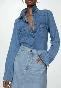 Camicia di jeans di un blu medio con colletto rilassato, due tasche frontali e bottoni; abbinata a una gonna di jeans azzurro chiaro.
