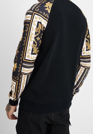 Person trägt schwarzen Pullover mit kunstvoll verzierten Ärmeln in Schwarz, Weiß und Gold, steht mit sichtbar Rücken und linker Seite.