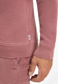 Rosa gestrickter Pullover mit strukturiertem Stoff, gerippten Bündchen und einem Logotag am Saum. Kombiniert mit rosa Cordhosen.