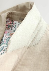 Col de manteau beige avec doublure en polaire blanche texturée et tissu intérieur imprimé floral dans des tons de rose, vert et crème.