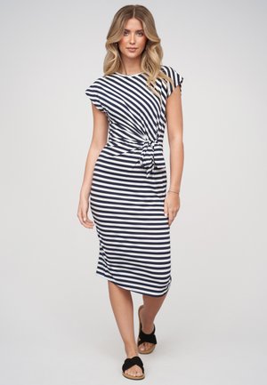 Donna con capelli biondi lunghi che indossa un vestito a righe blu navy e bianco fino al ginocchio con un nodo laterale e sandali neri che cammina verso avanti.
