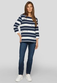 Frau mit langen braunen Haaren, die ein navy- und weißgestreiftes Shirt mit Text, blaue Jeans und weiße Sneakers trägt, steht vor einem grauen Hintergrund.