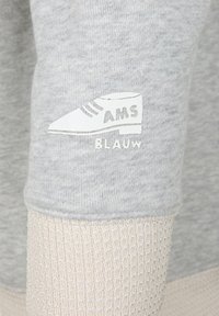 Tejido de sudadera gris con un puño de color crema texturizado; presenta un gráfico blanco de una zapatilla con el texto "AMS BLAUW."