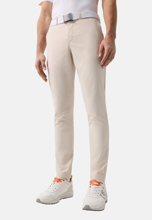 Mann trägt beigefarbene, schmal geschnittene Hose, weißen Gürtel mit Metallschnalle und weiße Turnschuhe mit orangen Schnürsenkeln, steht vor einem schlichten Hintergrund.