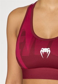 Tumši sarkans sporta krūšturis ar apaļu kakla izgriezumu, ribaina tekstūra, izteiksmīga logotipa detaļa un kontrastējošas tumšas svītras audumā.