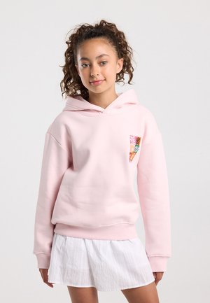 Mädchen mit lockigem Haar, das einen hellrosa Hoodie mit einem kleinen mehrfarbigen Aufnäher und einem weißen Rock trägt, steht vor einem einfarbigen Hintergrund.