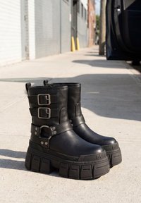 Paar schwarze klobige Plateaustiefel mit doppelten Schnallenriemen auf dem städtischen Gehweg neben einer geparkten Autotür.