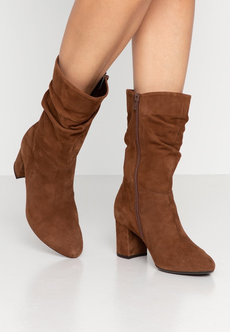 Bottes mi-mollet en daim marron avec talons carrés et fermetures éclair latérales portées par une personne debout sur une surface blanche réfléchissante.