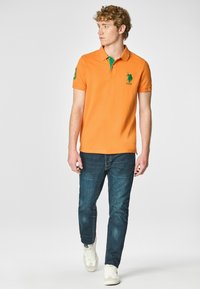 Polo arancione con colletto verde e logo, maniche corte, abbinato a jeans blu scuro e sneakers bianche. Sfondo semplice.