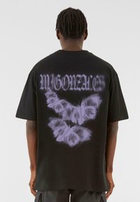 Tricou negru oversized cu un grafic mare de fluture lavandă și text stilizat "M GONZALEZ" pe spate. Mânici scurte și croială lejeră.