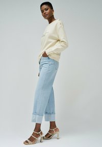 Sweatshirt couleur crème avec des accents de boutons sur l'épaule, associé à un jean large bleu clair et des talons hauts beiges à brides.