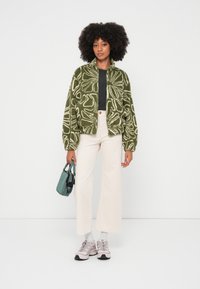 Chaqueta verde peluda con patrón abstracto blanco, top negro, pantalones anchos color crema y zapatillas rosas. Pequeño bolso verde con detalles en negro.