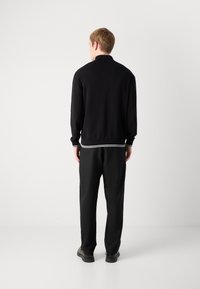 Μαύρο πλεκτό πουλόβερ με ριγωτό γιακά, μανσέτες και στρίφωμα. Συνδυάζεται με μαύρο παντελόνι και μινιμαλιστικά μαύρα παπούτσια. Εμφανίζεται από πίσω.