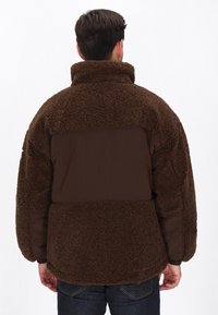 Mann, der eine dunkelbraune Fleecejacke mit strukturiertem Stoff und glatter Rückseite trägt, steht mit dem Rücken zu einer schlichten weißen Wand.