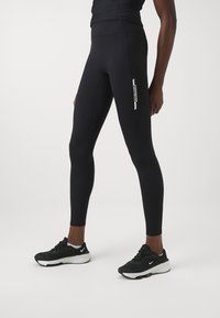 Fekete leggings rugalmas anyagból, testhezálló kialakítással. A combon egy fehér nyomtatott logó látható, sima, matt felülettel.