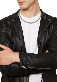 Sort læderbikerjakke med lynlås og trykknapkrave, lagdelt over en hvid t-shirt. Sølv kædehalskæde, arme krydset.