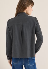 Dunkelgraues Hemd mit langen Ärmeln, Rückenschlitz-Detail und abgerundeten Manschetten. Getragen mit blauen Jeans, wobei der Fokus auf der Textur des Hemdes liegt.