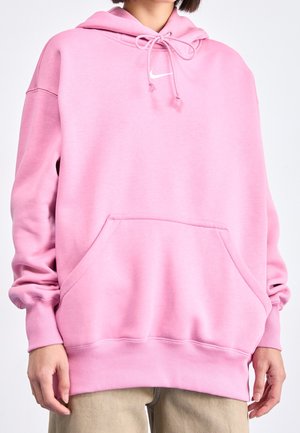 Personne portant un sweat à capuche Nike rose oversize avec poche devant et pantalon beige sur un fond uni.
