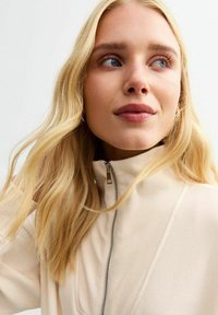 Giacca beige con zip e colletto alto, texture a coste e dettagli in metallo argentato, indossata da un individuo con lunghi capelli biondi mossi.