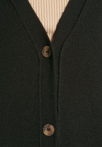 Czarny kardigan z dekoltem w serek, zapinany na dwa guziki, o gładkiej fakturze. Pod spodem częściowo widoczny jest beżowy, prążkowany element garderoby.