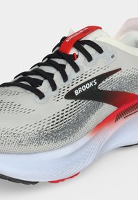 Scarpa da running Brooks grigio chiaro con lacci neri, dettagli rossi, tomaia in mesh traspirante e suola spessa bianca con dettaglio sfumato rosso.
