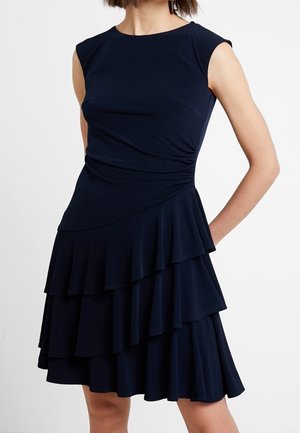 Robe bleu marine sans manches avec corsage ajusté et jupe à volants superposés, portée par une personne debout avec une main dans le dos.