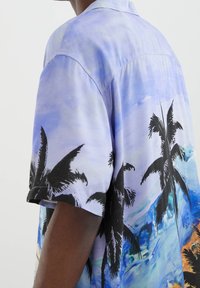 Persona que lleva una camisa de manga corta con un patrón de cielo azul y palmeras negras, mostrando la espalda y el brazo contra un fondo claro.
