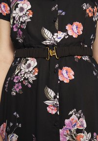 Musta kukkamekko, jossa on vaaleanpunaisia, purppuranvärisiä ja valkoisia kukkia, yksityiskohtina rypytetty musta vyö ja kultainen solki.