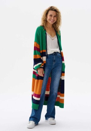 Uśmiechnięta kobieta ubrana w długi, paskowany kardigan, białą koszulkę na ramiączkach, niebieskie dżinsy i białe trampki, stojąca na tle jednolitego białego tła.