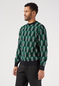 Maglione lavorato a maglia verde e nero con un intricato motivo ad arco e accenti bianchi. I polsini e l'orlo a coste offrono texture e vestibilità.