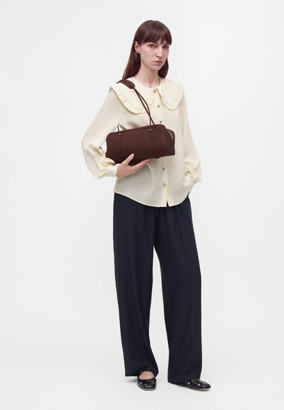 Femme portant un chemisier crème avec un col à volants et un pantalon noir à jambes larges tenant un sac à main marron avec des bretelles d'épaule.
