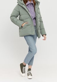 Lys grøn pufferjakke med hætte og lynlåslukning. Har quiltet struktur og forlommer, parret med lyseblå jeans og olivengrønne sneakers.