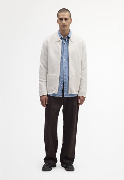 Hellbeige Wollmischung-Jacke mit zwei Fronttaschen, über ein blaues Denim-Hemd geschichtet, kombiniert mit braunen weiten Hosen und schwarzen Schuhen.