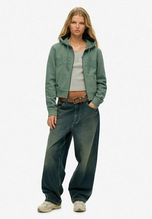 Sudadera verde recortada con bolsillos tipo canguro, top de tirantes gris ajustado y jeans anchos de color azul desgastado. Zapatillas completan el look informal.