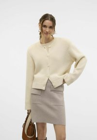 Cremefarbener gestrickter cardigan mit rundem Halsauschnitt, vorderen Knöpfen und geripptem Saum, kombiniert mit einem hellbeigen Rock mit vorderen Taschen.