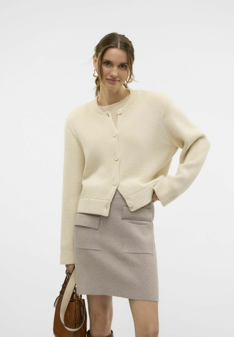 Cremefarbener gestrickter cardigan mit rundem Halsauschnitt, vorderen Knöpfen und geripptem Saum, kombiniert mit einem hellbeigen Rock mit vorderen Taschen.