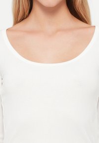 Γυναίκα φοράει μια λευκή ριγωτή μπλούζα με στρογγυλή λαιμόκοψη, που αποκαλύπτει τον λαιμό και την κλείδα, με μακριά ξανθά μαλλιά.